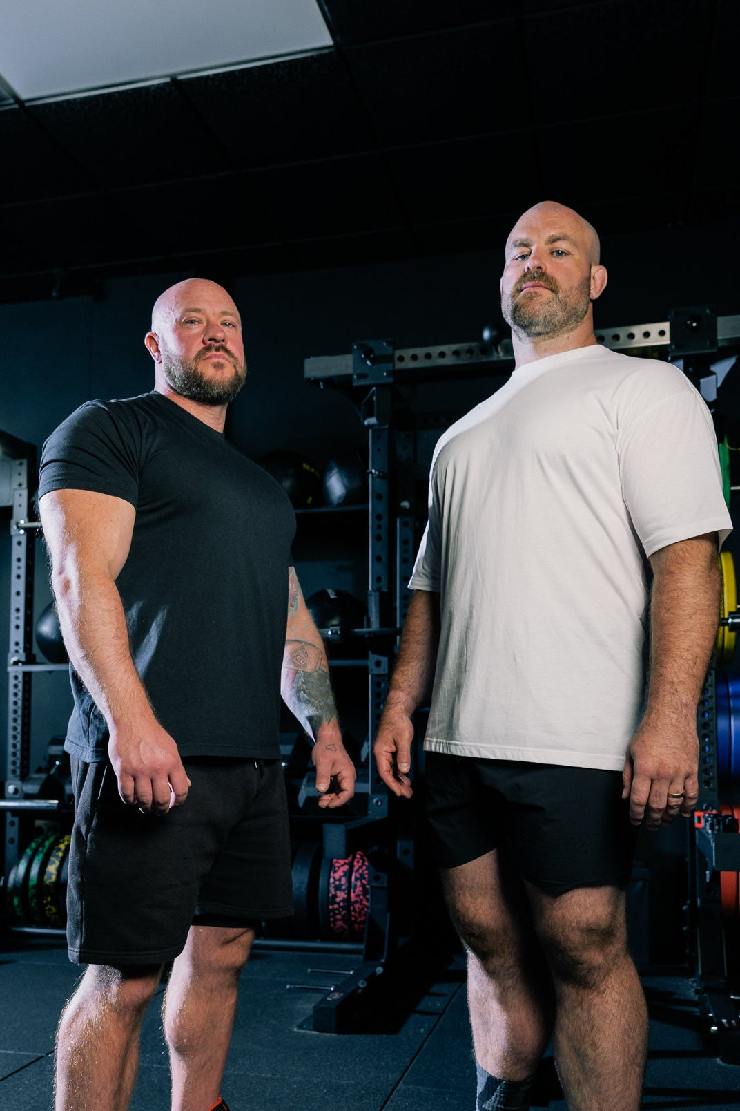 Load image into Gallery viewer, Two men standing in a dark gym environment, one wearing a black t-shirt and shorts with white sneakers, and the other wearing a white t-shirt and black shorts.
